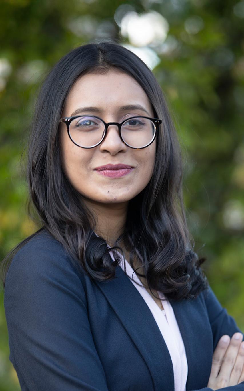 A woman smiling with confidence in front of a backdrop of greenery.